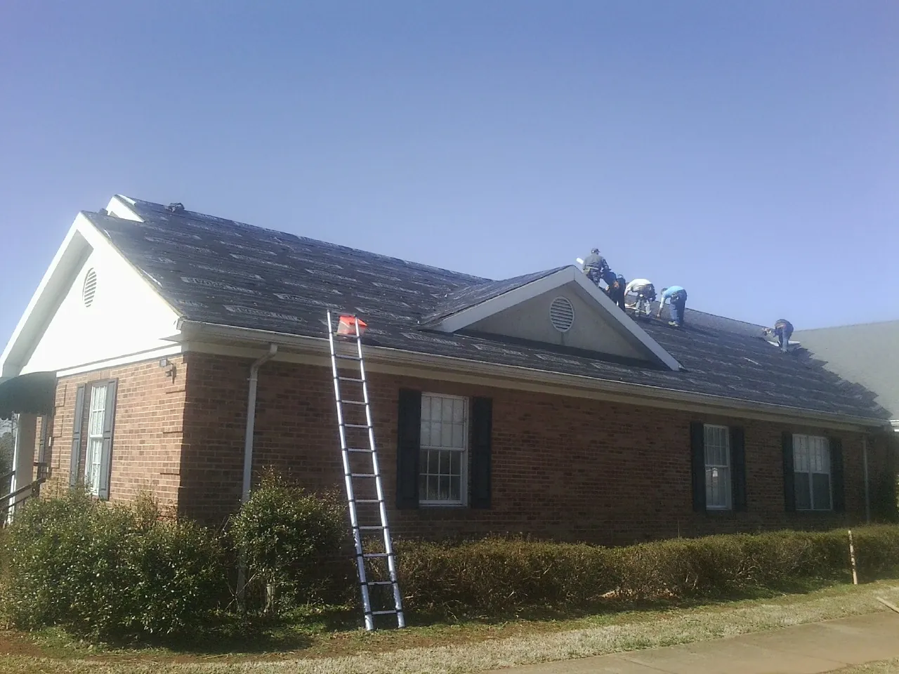 Crew tearing shingles off a brick ranch roof with a ladder set — Metal Roof Repair work by our crew in Wesley Chapel