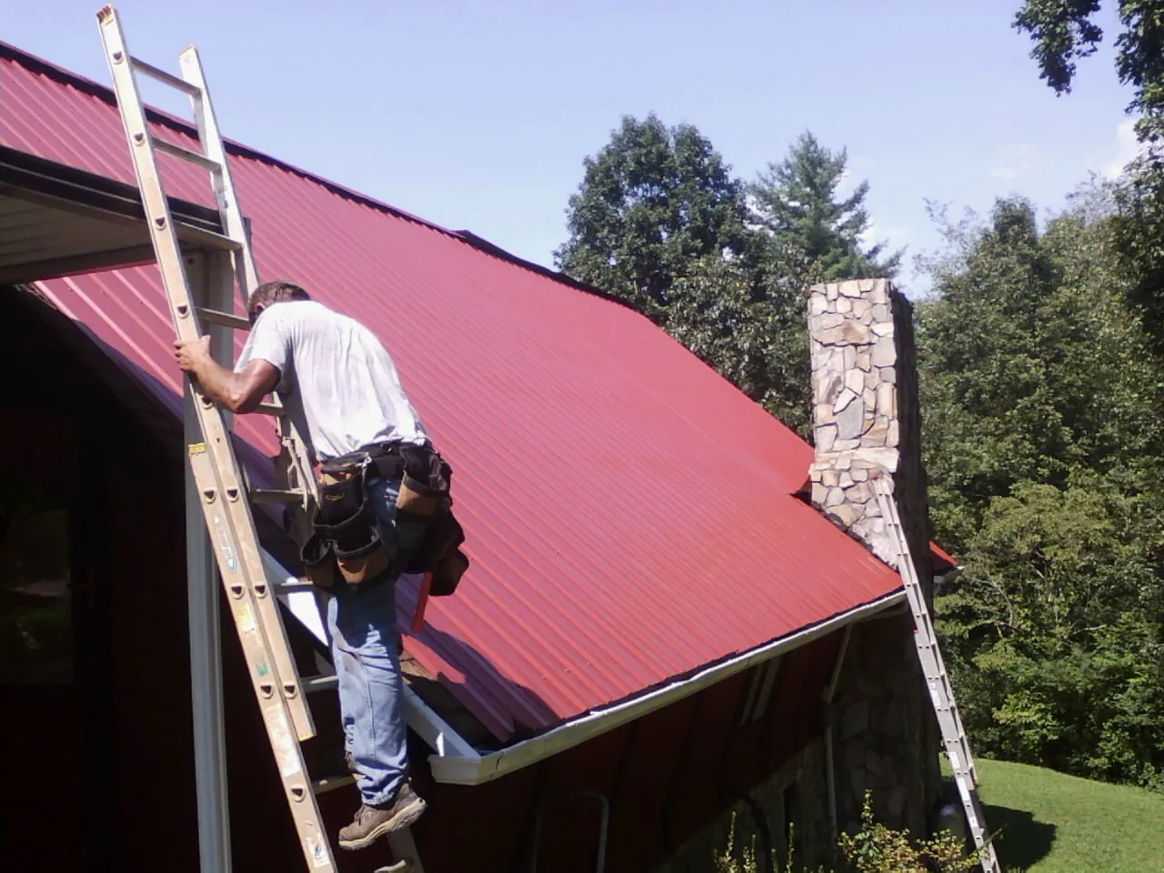 Roofer on a ladder beside a red metal roof and stone chimney — Soffit & Fascia Repair by Roofing Brothers in Wesley Chapel